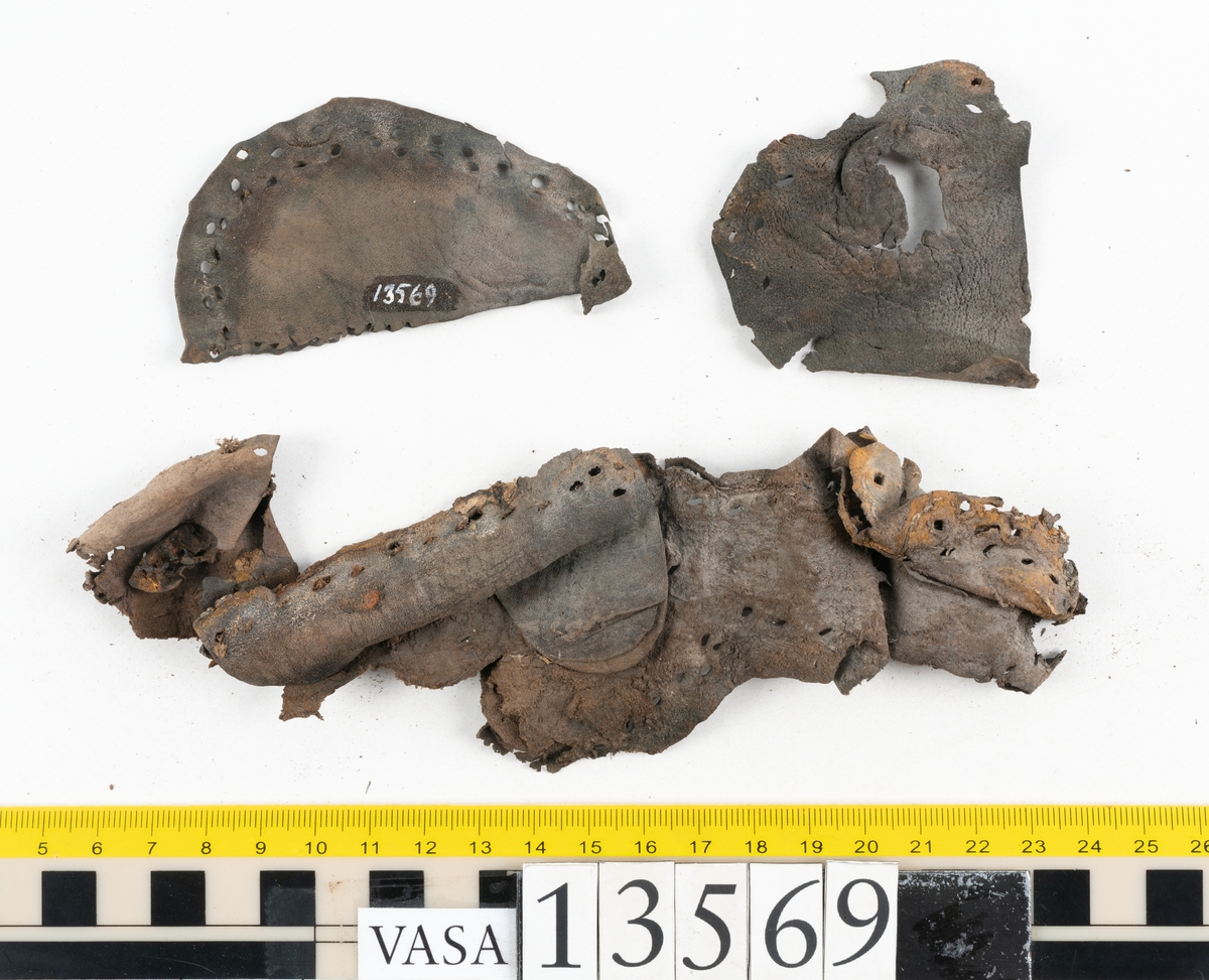 Ett 30-tal delar och fragment av sko: delar av inlägg, ovanläder, klack och sulor.

Ett ovanläder med kvarsittande tråd. Lädret är spaltat samt har rostliknande utfällningar. Två fragmentariska sulor med kvarsittande pligg. På flertalet fragment syns vita och rostliknande utfällningar.