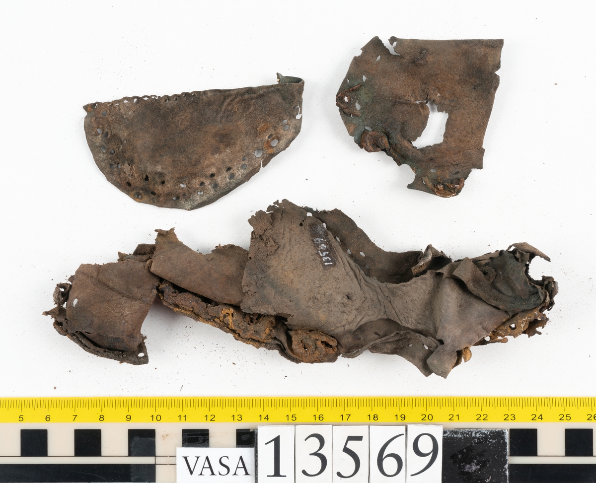 Ett 30-tal delar och fragment av sko: delar av inlägg, ovanläder, klack och sulor.

Ett ovanläder med kvarsittande tråd. Lädret är spaltat samt har rostliknande utfällningar. Två fragmentariska sulor med kvarsittande pligg. På flertalet fragment syns vita och rostliknande utfällningar.