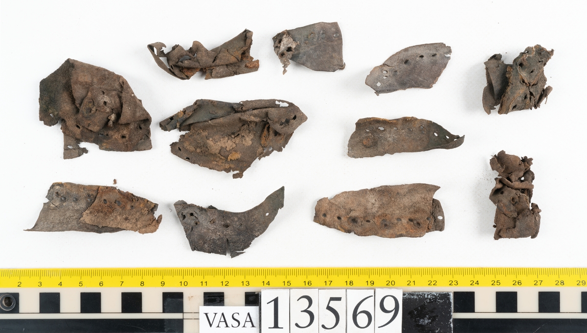 Ett 30-tal delar och fragment av sko: delar av inlägg, ovanläder, klack och sulor.

Ett ovanläder med kvarsittande tråd. Lädret är spaltat samt har rostliknande utfällningar. Två fragmentariska sulor med kvarsittande pligg. På flertalet fragment syns vita och rostliknande utfällningar.