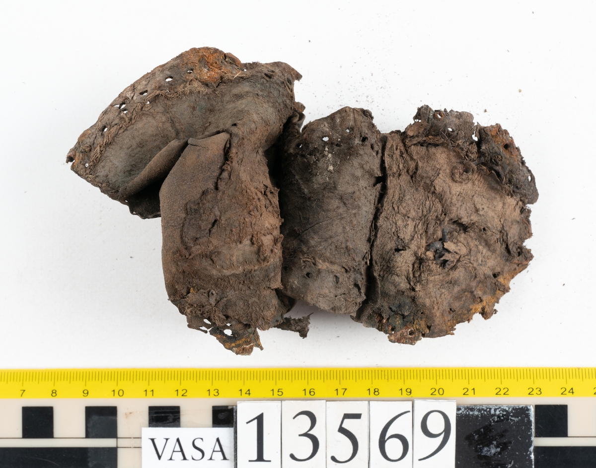 Ett 30-tal delar och fragment av sko: delar av inlägg, ovanläder, klack och sulor.

Ett ovanläder med kvarsittande tråd. Lädret är spaltat samt har rostliknande utfällningar. Två fragmentariska sulor med kvarsittande pligg. På flertalet fragment syns vita och rostliknande utfällningar.