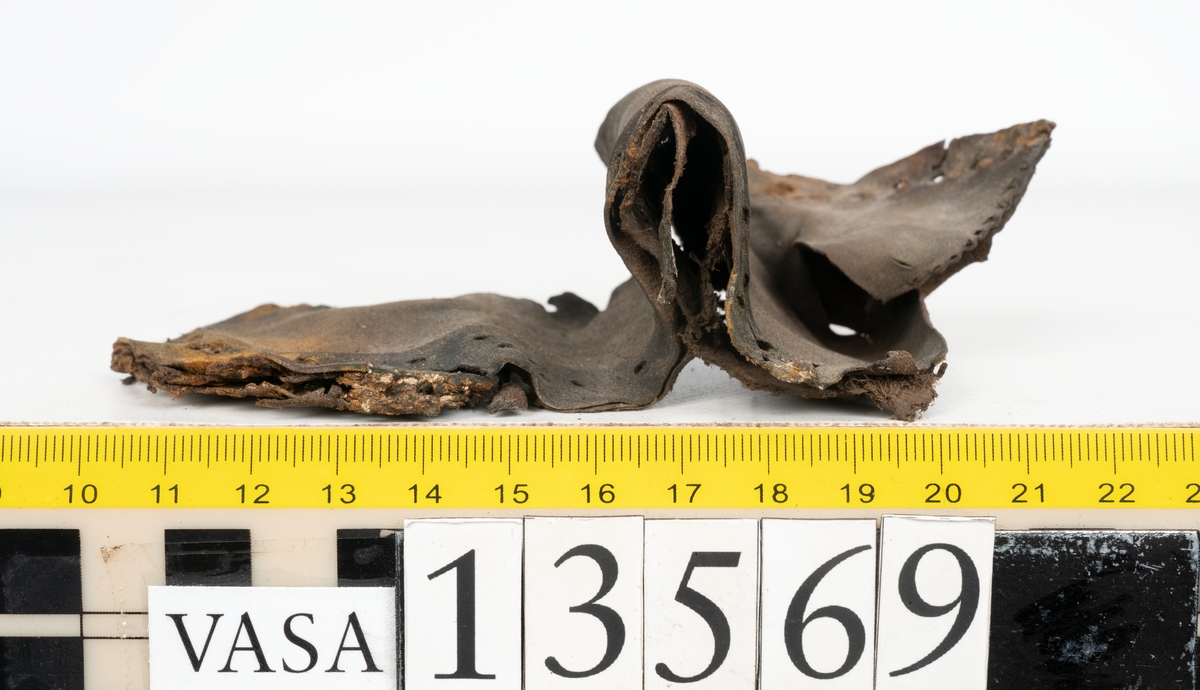 Ett 30-tal delar och fragment av sko: delar av inlägg, ovanläder, klack och sulor.

Ett ovanläder med kvarsittande tråd. Lädret är spaltat samt har rostliknande utfällningar. Två fragmentariska sulor med kvarsittande pligg. På flertalet fragment syns vita och rostliknande utfällningar.