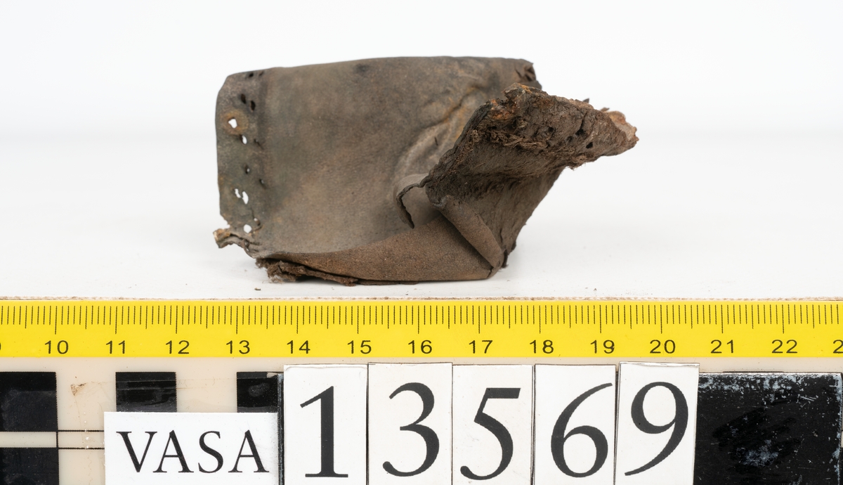 Ett 30-tal delar och fragment av sko: delar av inlägg, ovanläder, klack och sulor.

Ett ovanläder med kvarsittande tråd. Lädret är spaltat samt har rostliknande utfällningar. Två fragmentariska sulor med kvarsittande pligg. På flertalet fragment syns vita och rostliknande utfällningar.