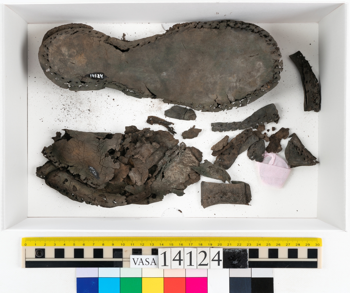 Sandalsydd högersko, troligtvis toffel, i delar.

Skobottnen består av två sulor med kvarsittanda skopligg och tråd. Vid tåpartiet sitter laglappar. På undersidan av yttersulan syns rostfärgade utfällningar.

Elva fragment från bottnen: sulor m m.

Ett fragmentariskt ovanläder. Ovanlädret har vita och rostfärgade utfällningar samt ligger hopvikt. Lädret är mycket sprött.
Totalt 15 fragment.