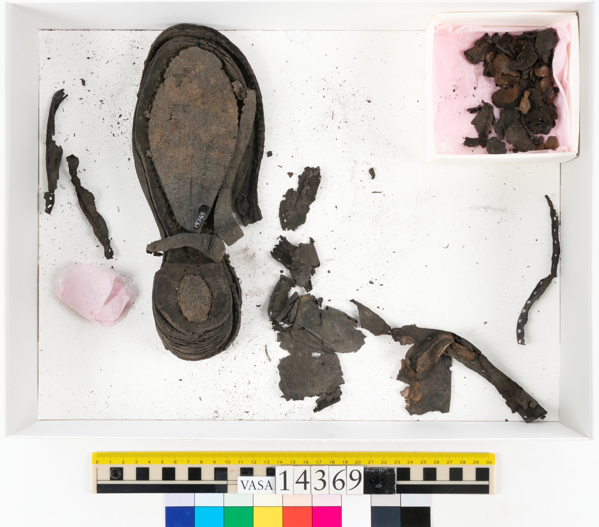 Delar och fragment från holtrandsydd sko: del av skobotten, två sulor samt ett 20-tal fragment från bottnen.

Skobottnen bestående av två sulor, rand och tre klacklappar. Bottnen har kvarsittande tråd och skopligg. Lädret är kraftigt.

En sula tillhörande skobottnen. Runt ytterkanten finns kvarsittande, grov, tråd. Lädret är kraftigt.  Det är något osäkert om den andra sulan hör till ovan skobotten.

Flertalet fragment har rostfärgade utfällningar.