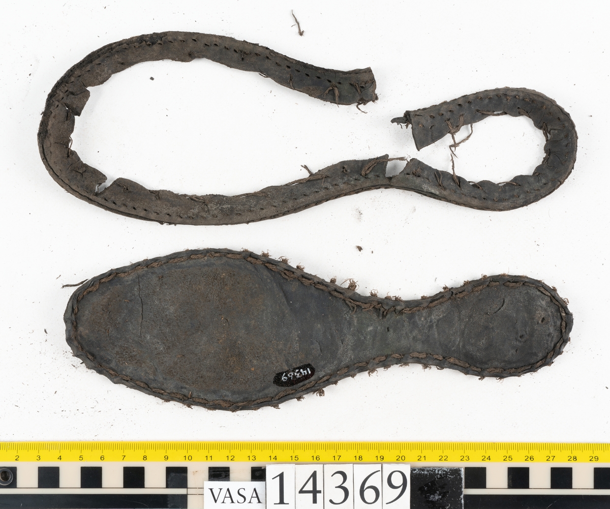 Delar och fragment från holtrandsydd sko: del av skobotten, två sulor samt ett 20-tal fragment från bottnen.

Skobottnen bestående av två sulor, rand och tre klacklappar. Bottnen har kvarsittande tråd och skopligg. Lädret är kraftigt.

En sula tillhörande skobottnen. Runt ytterkanten finns kvarsittande, grov, tråd. Lädret är kraftigt.  Det är något osäkert om den andra sulan hör till ovan skobotten.

Flertalet fragment har rostfärgade utfällningar.