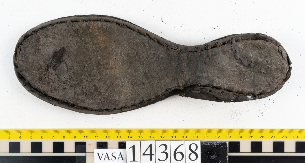 Dokumentation 1996:
Delar av sko: skobotten samt ett trettiotal fragment av ovanläder, holtrand m m.
Skobottnen består av tre sulor, holtrand och sex klacklappar. Mellan två av sulorna sitter klacklapparna fästade. En del av bakkappan sitter fästad vid bottnen.  Bottnen har kvarsittande tråd och vid hälpartiet finns kvarsittande skopligg.

Dokumentation 2022:
Skobotten till en randsko. Tillverkad i läder, med en läderklack. Skapades på en rak läst och bars på vänster fot. Inga dekorationer. Mittensulorna är delaminerade och är skurna längst med deras mittenpunkt. Randen är intakt och tå-delen är unik. 
För mer information, se Uppritning under Länkade filer.

Text in english:
This is the bottom unit of a shoe, made of leather, randed construction with a spring heel, made on a straight last and worn on the left foot, with no decorative features. The two midsoles are heavily delaminated and there is a slice along the centre of the sole. The rand is intact, and the toe wear is unique.
For more information, go to Länkade filer: Uppritning