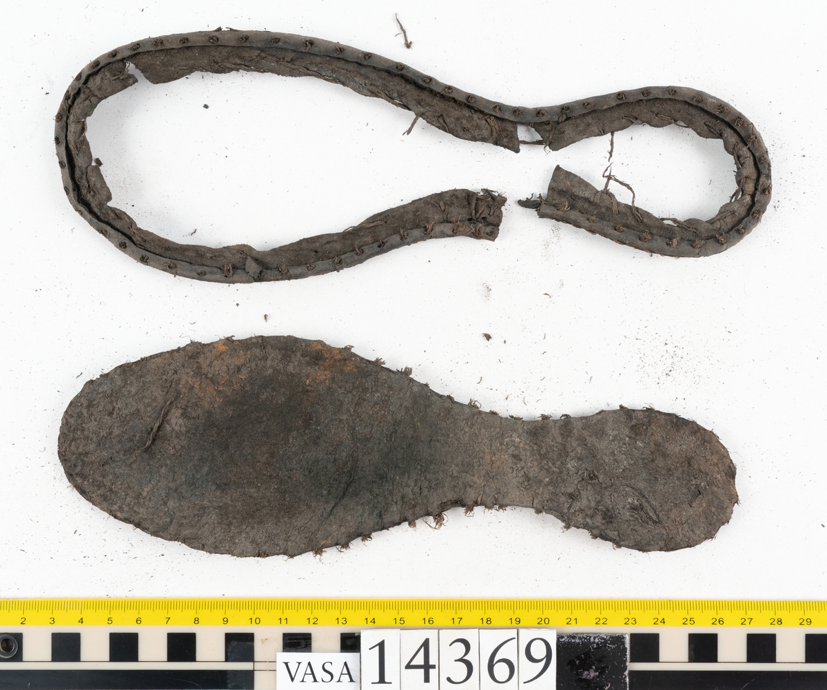 Delar och fragment från holtrandsydd sko: del av skobotten, två sulor samt ett 20-tal fragment från bottnen.

Skobottnen bestående av två sulor, rand och tre klacklappar. Bottnen har kvarsittande tråd och skopligg. Lädret är kraftigt.

En sula tillhörande skobottnen. Runt ytterkanten finns kvarsittande, grov, tråd. Lädret är kraftigt.  Det är något osäkert om den andra sulan hör till ovan skobotten.

Flertalet fragment har rostfärgade utfällningar.
