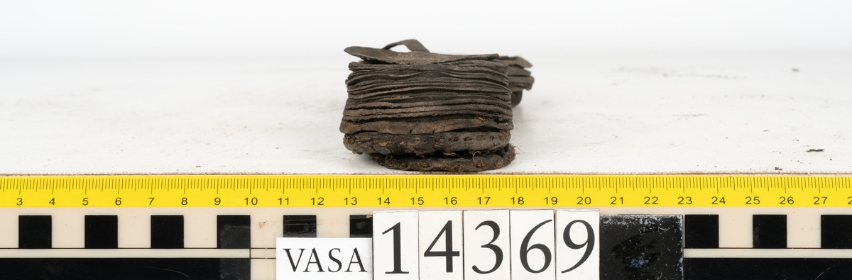Delar och fragment från holtrandsydd sko: del av skobotten, två sulor samt ett 20-tal fragment från bottnen.

Skobottnen bestående av två sulor, rand och tre klacklappar. Bottnen har kvarsittande tråd och skopligg. Lädret är kraftigt.

En sula tillhörande skobottnen. Runt ytterkanten finns kvarsittande, grov, tråd. Lädret är kraftigt.  Det är något osäkert om den andra sulan hör till ovan skobotten.

Flertalet fragment har rostfärgade utfällningar.
