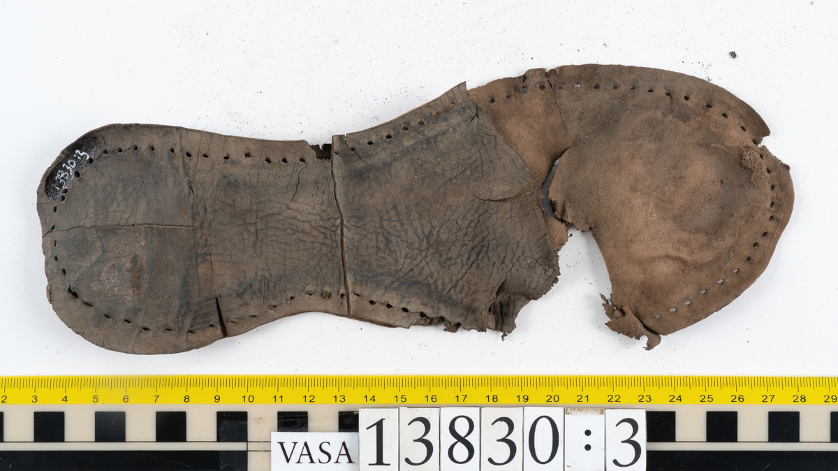 Dokumentation 1997:
Delar och fragment av sko: ovanläder, sulor, klacklappar, inlägg, holtrand, ett litet textilfragment mm.
Ett ovanläder, fyndnummer 13830. Ovanlädrets nedre del saknar narvsida. Ovanlädret är spaltat i två delar. Den avspaltade köttsidan har fyndnummer 13830:1.
Två stycken sulor, fyndnummer 13830:2 samt 13830:3. Sulornas tåparti respektive främre parti är fragmentariska.
Två sulor från höger skobotten, fyndnummer 13830:4 samt 13830:5. På sula fyndnummer 13830:5 finns kvarsittande skopligg.
En delvist fragmentarisk sula från vänster skobotten, fyndnummer 13830:6.
Ett 300-tal delar och fragment. Flertalet delar och fragment har hål efter träpligg och stygn på några delar finns kvarsittande tråd och/eller träpligg.

Dokumentation 2022:
Del av ovanläder. Framstycke till en stövel. Tillverkad som en svensksko. Plösen har en ovanlig form. Inga dekorationer. Lädret är delaminerat. 
För mer information, se Uppritning under Länkade filer.

Text in english:
This is a boot vamp with a tongue of a variant shape, made of leather of stitch-down construction, with no decorative features. The vamp has suffered delamination.
For more information, go to Länkade filer: Uppritning