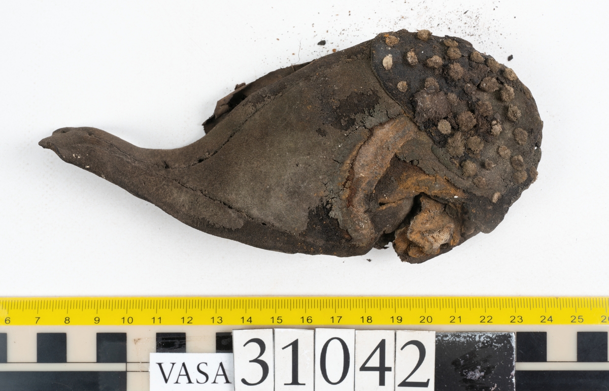 Del av en skobotten samt två fragmentariska sulor. Totalt ett tiotal fragment. Bottnens klackparti är fragmentariskt. På undersidan sitter halvmånformade laglappar. Kvarsittande skopligg. Lädret har rostfärgade utfällningar.