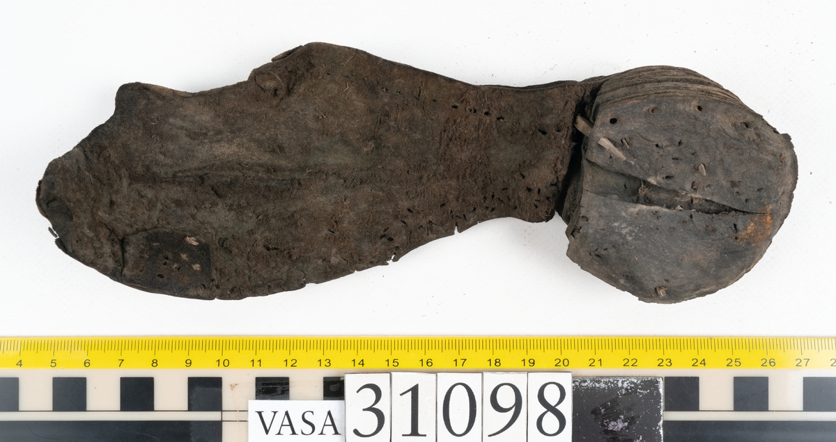 Dokumentation 1997:
Tre delar av sko: en del av en skobotten, en klacklapp och ett ovanläder samt två fragment som lossnat från dessa.
Del av skobotten i form av en yttersula med kvarsittande klack. Klacken är uppbyggd av 5-6 klacklappar. Sulans undersida är ritsad. Sulan är spaltad och tåpartiet är fragmentarisk. Sulan och klacken har kvarsittande träpligg.
En klacklapp med kvarsittande träpligg. Runt ytterkanterna och i klacklappens mitt finns hål efter träpligg.
Ett ovanläder samt en lös bit av plösen. På den lösa biten finns två snörhål. Längs med ovanlädrets ytterkanter löper stygnshål. Köttsidan har börjat att släppa från narvsidan.

Dokumentation 2022:
Del av ovanläder. Framstycke till en öppen svensksko som består av fragment. Tillverkad i läder. Inga dekorationer. Tillhörde en sko som bars på vänster fot. Ovanligt spetsig tå-del. Plösen är skadad och saknas. Det finns delaminering och veckning och inga tecken på lagningar. Det finns en tillhörande klack.
För mer information, se Länkade filer: Uppritning

Text in english:
This is the vamp of an open latchet shoe, made of leather, of stitch-down construction, worn on the left foot, with no decorative features, and an unusually pointed toe. The vamp consists of fragments, with the tongue broken off, and has suffered delamination and creasing. There are no signs of repairs. There is a heel associated with the vamp.
For more information, go to Länkade filer: Uppritning