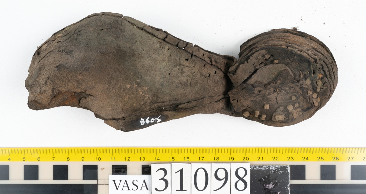 Dokumentation 1997:
Tre delar av sko: en del av en skobotten, en klacklapp och ett ovanläder samt två fragment som lossnat från dessa.
Del av skobotten i form av en yttersula med kvarsittande klack. Klacken är uppbyggd av 5-6 klacklappar. Sulans undersida är ritsad. Sulan är spaltad och tåpartiet är fragmentarisk. Sulan och klacken har kvarsittande träpligg.
En klacklapp med kvarsittande träpligg. Runt ytterkanterna och i klacklappens mitt finns hål efter träpligg.
Ett ovanläder samt en lös bit av plösen. På den lösa biten finns två snörhål. Längs med ovanlädrets ytterkanter löper stygnshål. Köttsidan har börjat att släppa från narvsidan.

Dokumentation 2022:
Del av ovanläder. Framstycke till en öppen svensksko som består av fragment. Tillverkad i läder. Inga dekorationer. Tillhörde en sko som bars på vänster fot. Ovanligt spetsig tå-del. Plösen är skadad och saknas. Det finns delaminering och veckning och inga tecken på lagningar. Det finns en tillhörande klack.
För mer information, se Länkade filer: Uppritning

Text in english:
This is the vamp of an open latchet shoe, made of leather, of stitch-down construction, worn on the left foot, with no decorative features, and an unusually pointed toe. The vamp consists of fragments, with the tongue broken off, and has suffered delamination and creasing. There are no signs of repairs. There is a heel associated with the vamp.
For more information, go to Länkade filer: Uppritning