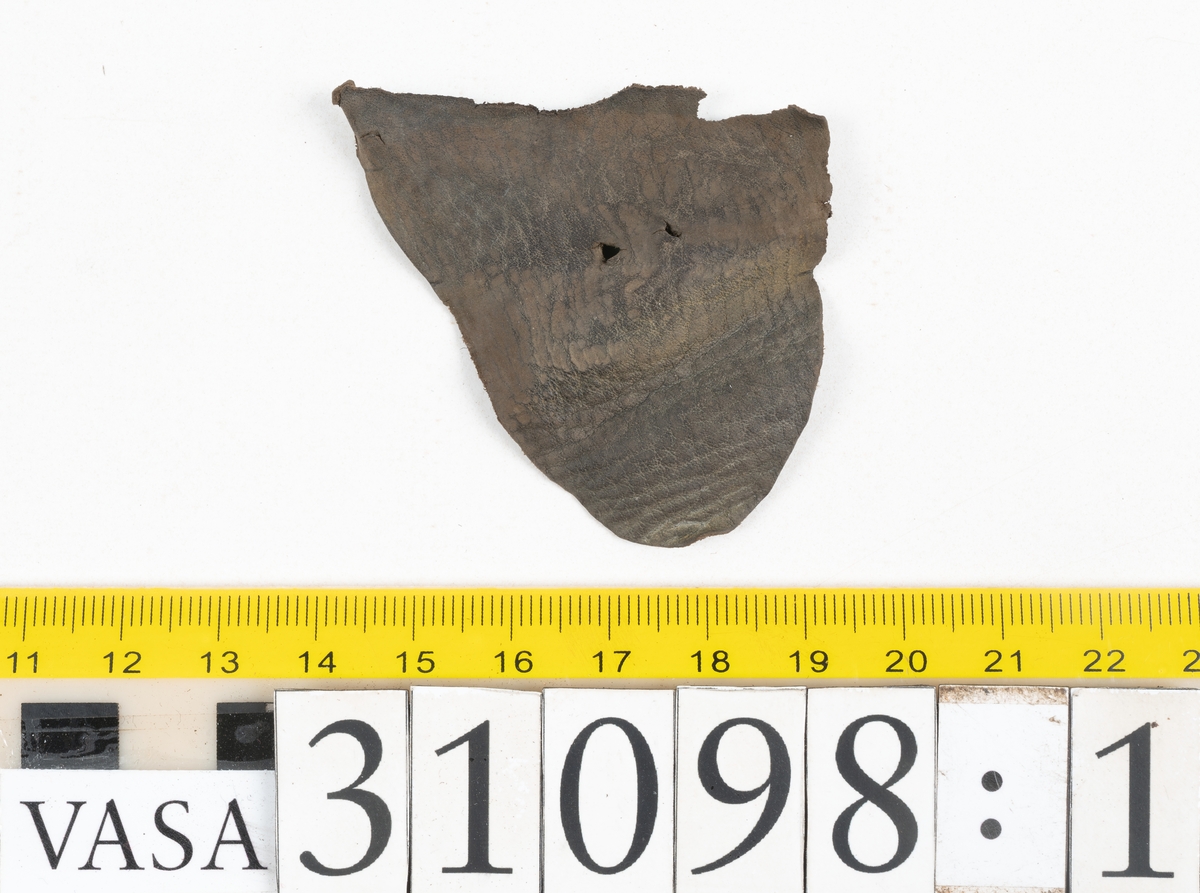 Dokumentation 1997:
Tre delar av sko: en del av en skobotten, en klacklapp och ett ovanläder samt två fragment som lossnat från dessa.
Del av skobotten i form av en yttersula med kvarsittande klack. Klacken är uppbyggd av 5-6 klacklappar. Sulans undersida är ritsad. Sulan är spaltad och tåpartiet är fragmentarisk. Sulan och klacken har kvarsittande träpligg.
En klacklapp med kvarsittande träpligg. Runt ytterkanterna och i klacklappens mitt finns hål efter träpligg.
Ett ovanläder samt en lös bit av plösen. På den lösa biten finns två snörhål. Längs med ovanlädrets ytterkanter löper stygnshål. Köttsidan har börjat att släppa från narvsidan.

Dokumentation 2022:
Del av ovanläder. Framstycke till en öppen svensksko som består av fragment. Tillverkad i läder. Inga dekorationer. Tillhörde en sko som bars på vänster fot. Ovanligt spetsig tå-del. Plösen är skadad och saknas. Det finns delaminering och veckning och inga tecken på lagningar. Det finns en tillhörande klack.
För mer information, se Länkade filer: Uppritning

Text in english:
This is the vamp of an open latchet shoe, made of leather, of stitch-down construction, worn on the left foot, with no decorative features, and an unusually pointed toe. The vamp consists of fragments, with the tongue broken off, and has suffered delamination and creasing. There are no signs of repairs. There is a heel associated with the vamp.
For more information, go to Länkade filer: Uppritning