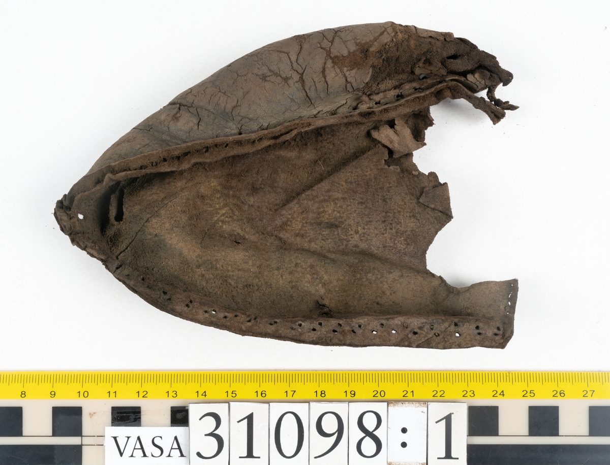 Dokumentation 1997:
Tre delar av sko: en del av en skobotten, en klacklapp och ett ovanläder samt två fragment som lossnat från dessa.
Del av skobotten i form av en yttersula med kvarsittande klack. Klacken är uppbyggd av 5-6 klacklappar. Sulans undersida är ritsad. Sulan är spaltad och tåpartiet är fragmentarisk. Sulan och klacken har kvarsittande träpligg.
En klacklapp med kvarsittande träpligg. Runt ytterkanterna och i klacklappens mitt finns hål efter träpligg.
Ett ovanläder samt en lös bit av plösen. På den lösa biten finns två snörhål. Längs med ovanlädrets ytterkanter löper stygnshål. Köttsidan har börjat att släppa från narvsidan.

Dokumentation 2022:
Del av ovanläder. Framstycke till en öppen svensksko som består av fragment. Tillverkad i läder. Inga dekorationer. Tillhörde en sko som bars på vänster fot. Ovanligt spetsig tå-del. Plösen är skadad och saknas. Det finns delaminering och veckning och inga tecken på lagningar. Det finns en tillhörande klack.
För mer information, se Länkade filer: Uppritning

Text in english:
This is the vamp of an open latchet shoe, made of leather, of stitch-down construction, worn on the left foot, with no decorative features, and an unusually pointed toe. The vamp consists of fragments, with the tongue broken off, and has suffered delamination and creasing. There are no signs of repairs. There is a heel associated with the vamp.
For more information, go to Länkade filer: Uppritning