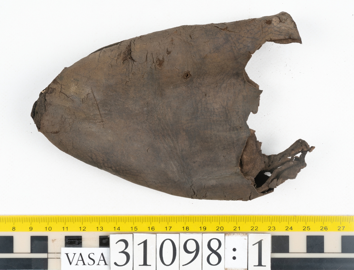 Dokumentation 1997:
Tre delar av sko: en del av en skobotten, en klacklapp och ett ovanläder samt två fragment som lossnat från dessa.
Del av skobotten i form av en yttersula med kvarsittande klack. Klacken är uppbyggd av 5-6 klacklappar. Sulans undersida är ritsad. Sulan är spaltad och tåpartiet är fragmentarisk. Sulan och klacken har kvarsittande träpligg.
En klacklapp med kvarsittande träpligg. Runt ytterkanterna och i klacklappens mitt finns hål efter träpligg.
Ett ovanläder samt en lös bit av plösen. På den lösa biten finns två snörhål. Längs med ovanlädrets ytterkanter löper stygnshål. Köttsidan har börjat att släppa från narvsidan.

Dokumentation 2022:
Del av ovanläder. Framstycke till en öppen svensksko som består av fragment. Tillverkad i läder. Inga dekorationer. Tillhörde en sko som bars på vänster fot. Ovanligt spetsig tå-del. Plösen är skadad och saknas. Det finns delaminering och veckning och inga tecken på lagningar. Det finns en tillhörande klack.
För mer information, se Länkade filer: Uppritning

Text in english:
This is the vamp of an open latchet shoe, made of leather, of stitch-down construction, worn on the left foot, with no decorative features, and an unusually pointed toe. The vamp consists of fragments, with the tongue broken off, and has suffered delamination and creasing. There are no signs of repairs. There is a heel associated with the vamp.
For more information, go to Länkade filer: Uppritning