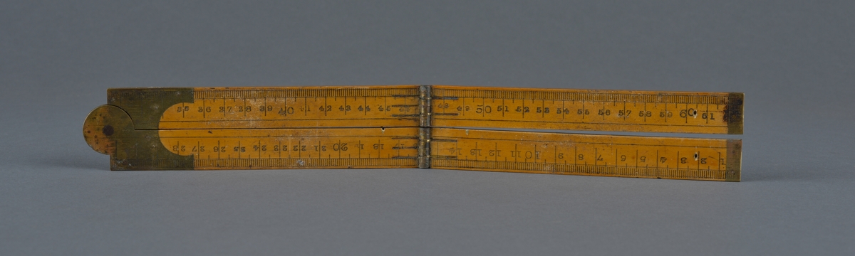 Lengdemål, sammenslåbar. 
Et hjul i midten og to ledd gjør at måleredskapet enkelt kan slås sammen og puttes i lommen. 

Laget i tre og metall.
Centimetermål på den ene siden og tommer på den andre.