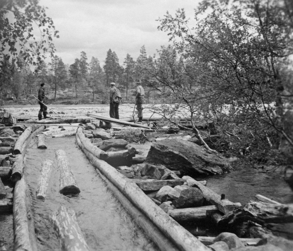 Fra den såkalte Feragkanalen, ei vannrenne mellom innsjøene Femund og Feragen, som ligger cirka 8 meter lavere. De første spadestikkene for å få til en kanal på denne strekningen skal ha vært tatt alt i 1714, men kanalen var ikke klar for tømmerfløting før i 1764. Feragkanalen ble bygd for Røros kobberverk, som stadig stevde med knapphet på energivirke. Dansknorske myndigheter lot verket få bruke virke fra statsallmenningene rundt Femund for å sikre stabil drift i kobberproduksjonen i verkets smeltehytter, og dermed skatteinntekter til staten. Kobberverkets behov for kanalen avtok etter at Røros i 1877 gjorde det mulig for verket og fyre med steinkull i stedet for trekull. I åra som fulgte ble det ført rettsaker der staten vant tilbake disposisjonsretten til de utmarksarealene rundt Femund. Fra da av var det andre instanser som transporterte tømmer gjennom kanalen, som skal ha vært i bruk på denne måten helt fram til 1973. Dette fotografiet må være tatt i denne seine fasen. Vi ser tre menn, hvorav to hadde fløterhaker som de brukte til å hake tømmer inn i renna med. Akkurat her var renna var lagd av rundtømmer som fundert steinfylte tømmerkasser.