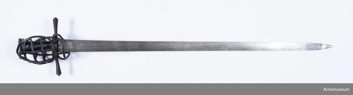 Klingan är tveeggad, har upptill på bägge sidor grund skålslipning eller bred, grund blodrand, men är i övrigt flack. Fästet är av svärtat järn och har hel gallerkorg. Knappen är låg och har upptill formen av en stympad, fyrkantig pyramid. Där- efter följer nedåt en flat 3 mm bred kant. Knappens nedre del  bildar en slags kort, bred, nedåt smalare hals med ovalt tvärsnitt. Kaveln har ovalt tvärsnitt, är vidare uppåt och är lindad med snören, som bestrukits med svart fernissa. Parerstången är rak, har kvadratiskt tvärsnitt med en kant vänd uppåt. Mot ändarna vidgas den, men ändarna har pyramidformig avslutning. Handbygelns övre ända är framåtböjd och står nära och i jämnhöjd med knappen. Framtill utgår från korset en fingerhake. På terssidan finns vid korset en stor, oval parerring. Från fingerhakens nedre ända går på terssidan en spång utåt och uppåt till parerringen, vilken den träffar strax framför dess bakre krök. Från motsvarande ställe på parerringens  övre sida går en spång något bakåt sedan utåt och uppåt, så inåt och litet nedåt till handbygelns övre del, som den träffar strax nedanför bygelns övre ända. Från den horisontala parerbygelns mitt och strax bakom dess främre krök går från vardera stället en bygel i båge uppåt till den nyss omtalade tersspången, vilken dessa byglar träffar i samma punkt ett stycke bakom dess övre ända. I övrigt utgörs skyddet på terssidan av tre horisontala spänger, som går från handbygeln till tersspången, och som tillsammans har formen av ett på bredden starkt utdraget S. På kvartsidan går från fingerhakens nedre ända en spång till parerstångens bakre arm, som den träffar strax bakom korset. Från mitten av denna spång går en annan längre spång något bakåt, sedan utåt och uppåt, så framåt och slutligen nedåt till handbygeln, som den träffar strax nedanför bygelns övre ända. Strax bakom den främre kröken på den förstnämnda kvartspången går en bygel i båge uppåt till den sistnämnda spången, som den träffar en bit bakom dess övre ända. I övrigt utgörs skyddet på kvartsidan av tre horisontala spänger, som går från handbygeln till den längre kvartspången. Dessa tre sistnämnda kvartspänger har tillsammans formen av ett på bredden starkt utdraget S. Alla byglar och spänger har cirkelrunt tvärsnitt.