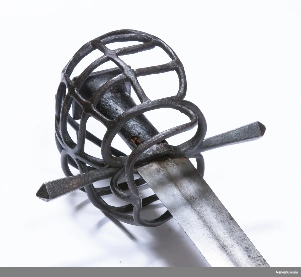 Klingan är tveeggad, har upptill på bägge sidor grund skålslipning eller bred, grund blodrand, men är i övrigt flack. Fästet är av svärtat järn och har hel gallerkorg. Knappen är låg och har upptill formen av en stympad, fyrkantig pyramid. Där- efter följer nedåt en flat 3 mm bred kant. Knappens nedre del  bildar en slags kort, bred, nedåt smalare hals med ovalt tvärsnitt. Kaveln har ovalt tvärsnitt, är vidare uppåt och är lindad med snören, som bestrukits med svart fernissa. Parerstången är rak, har kvadratiskt tvärsnitt med en kant vänd uppåt. Mot ändarna vidgas den, men ändarna har pyramidformig avslutning. Handbygelns övre ända är framåtböjd och står nära och i jämnhöjd med knappen. Framtill utgår från korset en fingerhake. På terssidan finns vid korset en stor, oval parerring. Från fingerhakens nedre ända går på terssidan en spång utåt och uppåt till parerringen, vilken den träffar strax framför dess bakre krök. Från motsvarande ställe på parerringens  övre sida går en spång något bakåt sedan utåt och uppåt, så inåt och litet nedåt till handbygelns övre del, som den träffar strax nedanför bygelns övre ända. Från den horisontala parerbygelns mitt och strax bakom dess främre krök går från vardera stället en bygel i båge uppåt till den nyss omtalade tersspången, vilken dessa byglar träffar i samma punkt ett stycke bakom dess övre ända. I övrigt utgörs skyddet på terssidan av tre horisontala spänger, som går från handbygeln till tersspången, och som tillsammans har formen av ett på bredden starkt utdraget S. På kvartsidan går från fingerhakens nedre ända en spång till parerstångens bakre arm, som den träffar strax bakom korset. Från mitten av denna spång går en annan längre spång något bakåt, sedan utåt och uppåt, så framåt och slutligen nedåt till handbygeln, som den träffar strax nedanför bygelns övre ända. Strax bakom den främre kröken på den förstnämnda kvartspången går en bygel i båge uppåt till den sistnämnda spången, som den träffar en bit bakom dess övre ända. I övrigt utgörs skyddet på kvartsidan av tre horisontala spänger, som går från handbygeln till den längre kvartspången. Dessa tre sistnämnda kvartspänger har tillsammans formen av ett på bredden starkt utdraget S. Alla byglar och spänger har cirkelrunt tvärsnitt.