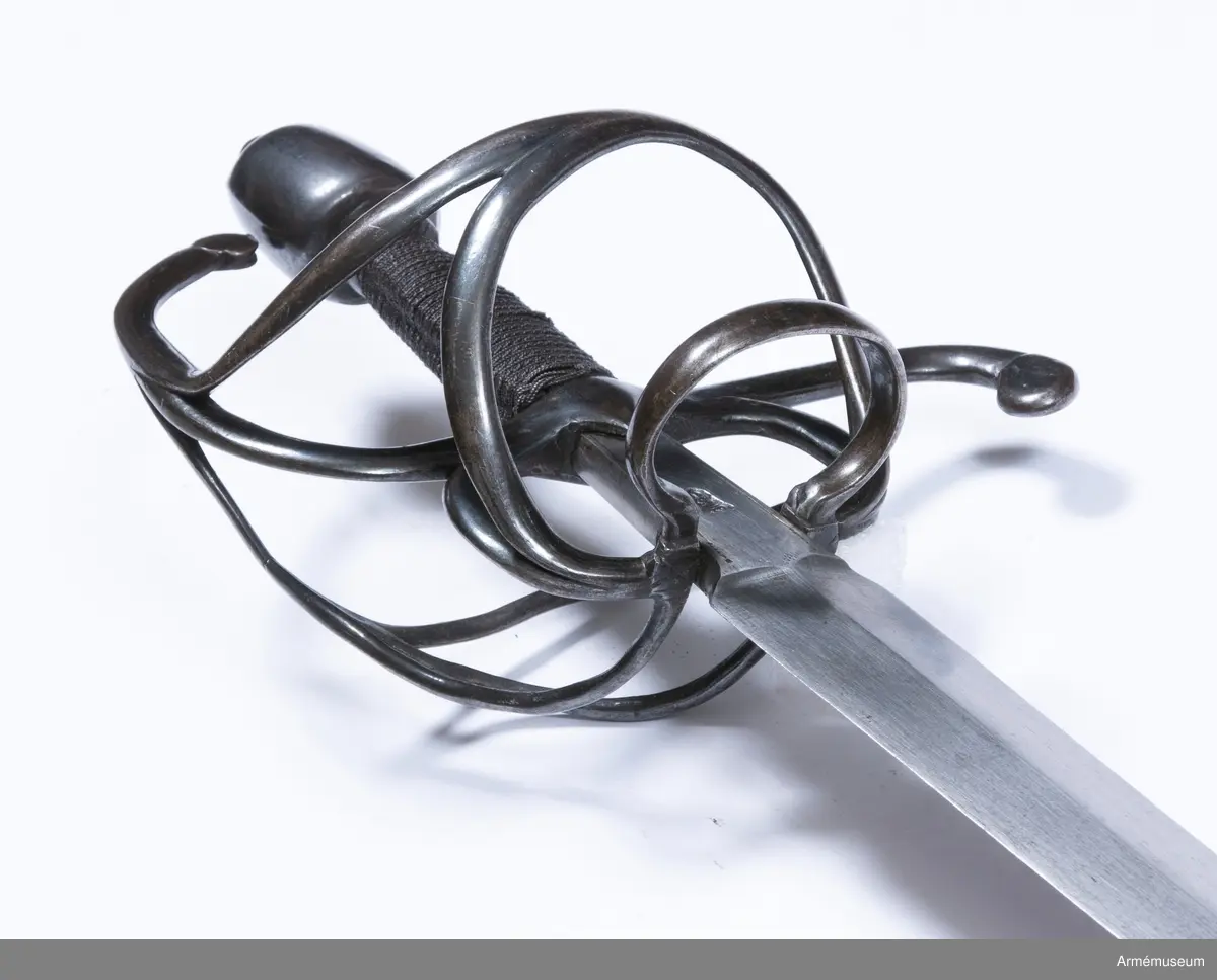 Den tveeggade klingan är upptill rätt bred samt avsmalnar långsamt och likformigt mot udden så att den får formen av en långdragen triangel. På vardera flatsidan har klingan längsgående rygg, och upptill en cirka 65 mm lång ansats, som har plana flatsidor och svagt kullriga eggsidor. Vid klingbasen är ansatsen 28 mm men vid fästet 19 mm bred. Längs var och en av ansatsens långkanter går på flatsidorna en ingraverad rand. På var och en av ansatsens flatsidor finns en krönt stämpel med bokstaven S över bokstaven T, samt på innersidan dessutom bokstäverna H Z under en krona, vilket lär betyda Hauptzeughaus. Fästet är av svärtat järn. Knappen har närmast formen av en långdragen, stympad kon med kullrig översida och med rygg längs var eggsida, så att den får spetsovalt tvärsnitt.  Nitknappen är rätt hög och avskiljes genom en horisontal fals  från knappen. Knappens hals är kort och kullrig samt  avskiljes även den genom en horisontal fals från knappen. På yttersidan av knapphalsens underkant finns fyra inhuggna streck.

Träkaveln är närmast spolformig, men betydligt bredare vid korset än vid knappen. Flatsidorna är kullriga, men längs vardera eggsidan går en försänkt rand. Nedtill har kaveln krans av snodd järntråd, men den övre kransen är förkommen. Kavellindningen består av en rätt grov, överspunnen järntråd samt av en höger- och en vänstersnodd järntråd, vardera hoptvinnad av två parter. Parerstången har spetsovalt tvärsnitt samt vid korset på var sida en trekantig, nedåtvänd flik. Baktill är parerstången nedböjd och avslutas med en skivformig utsvällning, som är svängd litet åt yttersidan. Framtill övergår parerstången bågformigt i handbygeln, vilken upptill avslutas med en flat skiva, vars kant står några millimeter framför knappen. Från korset utgår vid var och en av ansatsens eggsidor en stor,  nedåtriktad fingerhake. De båda fingerhakarnas nedre ändar förenas på yttersidan med en horisontal bygel. På samma sida utgår även en från fingerhakarnas nedre ändar en snett uppåtriktad, i ca 45 graders vinkel ställd stor parerbygel, vars övre kant står i jämnhöjd med kavelns nedre del. Från  den hösta punkten på denna bygel går en spång till handbygelns mitt. På innersidan går en nedtill tregrenigt S-formig gaffelspång från handbygelns mitt till fingerhakarnas nedre ändar. Gaffelspångens olika delar har cirkelrunt tvärsnitt, men yttersidans byglar och spång har kullrig utsida och flat insida.