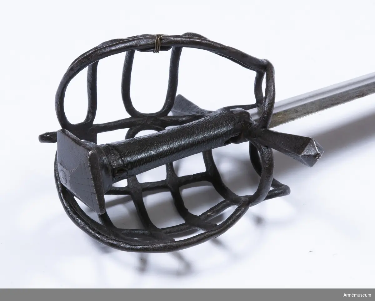 Klingan är tveeggad, har upptill på bägge sidor grund skålslipning eller bred, grund blodrand, men är i övrigt flack. Fästet är av svärtat järn och har hel gallerkorg. Knappen är låg och har upptill formen av en stympad, fyrkantig pyramid. Där- efter följer nedåt en flat 3 mm bred kant. Knappens nedre del  bildar en slags kort, bred, nedåt smalare hals med ovalt tvärsnitt. Kaveln har ovalt tvärsnitt, är vidare uppåt och är lindad med snören, som bestrukits med svart fernissa. Parerstången är rak, har kvadratiskt tvärsnitt med en kant vänd uppåt. Mot ändarna vidgas den, men ändarna har pyramidformig avslutning. Handbygelns övre ända är framåtböjd och står nära och i jämnhöjd med knappen. Framtill utgår från korset en fingerhake. På terssidan finns vid korset en stor, oval parerring. Från fingerhakens nedre ända går på terssidan en spång utåt och uppåt till parerringen, vilken den träffar strax framför dess bakre krök. Från motsvarande ställe på parerringens  övre sida går en spång något bakåt sedan utåt och uppåt, så inåt och litet nedåt till handbygelns övre del, som den träffar strax nedanför bygelns övre ända. Från den horisontala parerbygelns mitt och strax bakom dess främre krök går från vardera stället en bygel i båge uppåt till den nyss omtalade tersspången, vilken dessa byglar träffar i samma punkt ett stycke bakom dess övre ända. I övrigt utgörs skyddet på terssidan av tre horisontala spänger, som går från handbygeln till tersspången, och som tillsammans har formen av ett på bredden starkt utdraget S. På kvartsidan går från fingerhakens nedre ända en spång till parerstångens bakre arm, som den träffar strax bakom korset. Från mitten av denna spång går en annan längre spång något bakåt, sedan utåt och uppåt, så framåt och slutligen nedåt till handbygeln, som den träffar strax nedanför bygelns övre ända. Strax bakom den främre kröken på den förstnämnda kvartspången går en bygel i båge uppåt till den sistnämnda spången, som den träffar en bit bakom dess övre ända. I övrigt utgörs skyddet på kvartsidan av tre horisontala spänger, som går från handbygeln till den längre kvartspången. Dessa tre sistnämnda kvartspänger har tillsammans formen av ett på bredden starkt utdraget S. Alla byglar och spänger har cirkelrunt tvärsnitt.