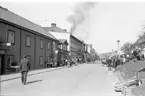 Det brinner i en fastighet utmed Brahegatan i Gränna. De ryker från taket. Brandmän står på gatan och bekämpar elden. En folksamling på trottoaren mittemot.