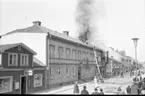 Det brinner i en fastighet utmed Brahegatan i Gränna. De ryker från taket där brandmän befinner sig. En folksamling på trottoaren mittemot.