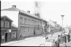 Det brinner i en fastighet utmed Brahegatan i Gränna. De ryker från taket där brandmän befinner sig. En folksamling på trottoaren mittemot.