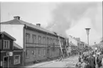 Det brinner i en fastighet utmed Brahegatan i Gränna. De ryker från taket där brandmän befinner sig. En folksamling på trottoaren mittemot.