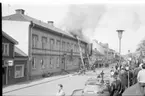 Det brinner i en fastighet utmed Brahegatan i Gränna. De ryker från taket där brandmän befinner sig. En folksamling på trottoaren mittemot.