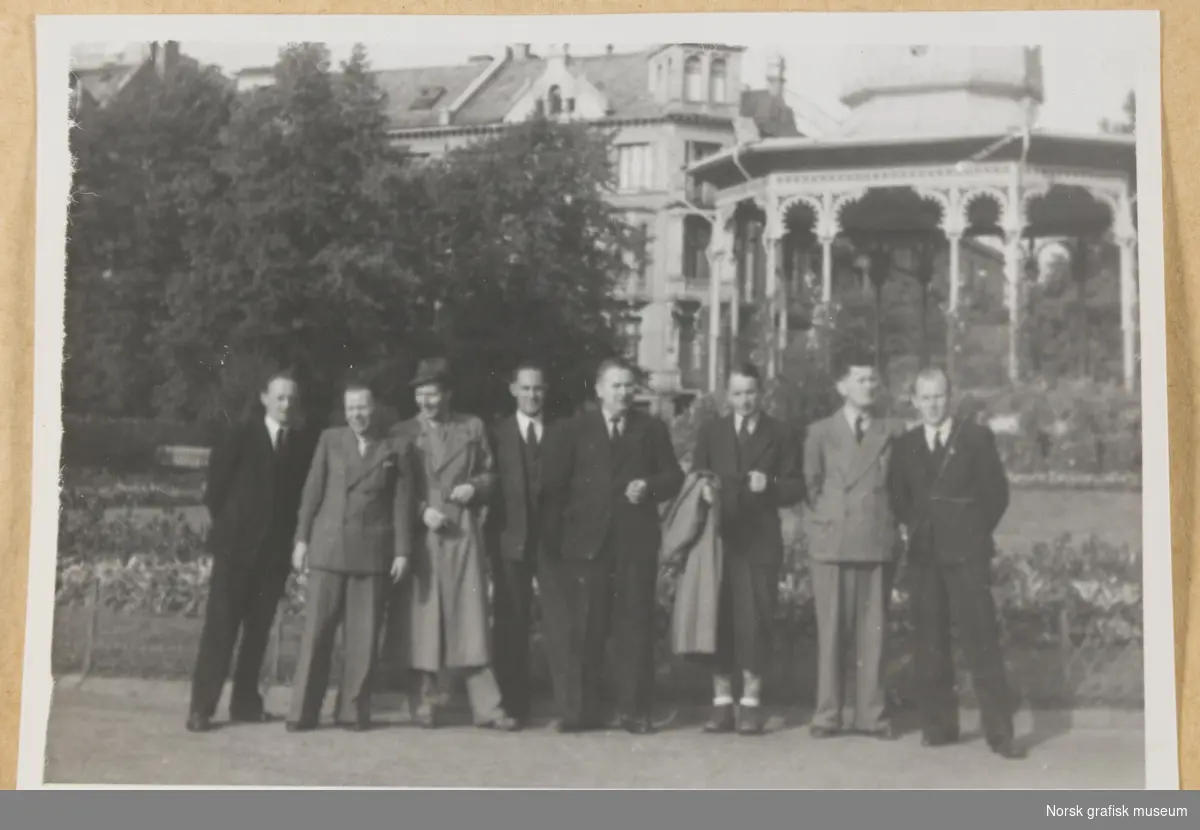 Utendørs gruppebilde. Åtte dresskledde menn står i Byparken med Musikkpaviljongen i bakgrunnen. Fotografert i forbindelse med Vestlandsk Trykkerstevne i Bergen, 1946.