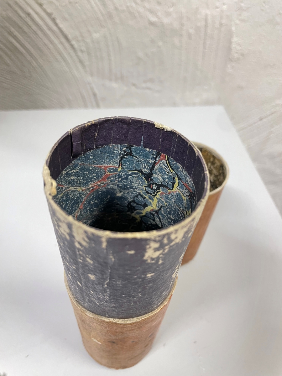 Cylinderformat fodral av papp, klädd med gulbrunt papper. Kanterna vid öppningen har förgyllda bårder. Ena kortänden har initialerna E. T. i förgyllning. Inuti ligger en kort fjäderpenna.