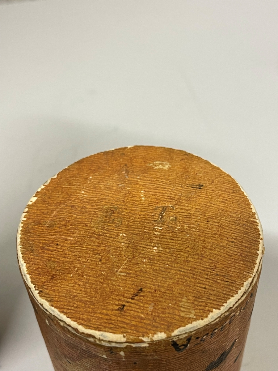 Cylinderformat fodral av papp, klädd med gulbrunt papper. Kanterna vid öppningen har förgyllda bårder. Ena kortänden har initialerna E. T. i förgyllning. Inuti ligger en kort fjäderpenna.