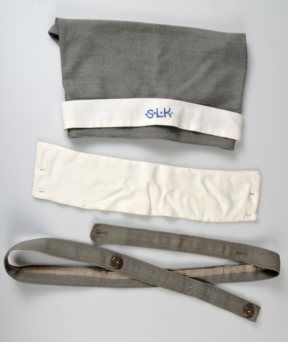 Klänning av grått ylletyg. Knälång klänning med isydd lång ärm. Kjolen har två nedsydda vecka fram och två fickor och ett veck bak. Knäppning från krage till midja med tio grå knappar.Knappar i handled.  Påsydda axelklaffar. Manchetter i vitt, löstagbara, påknäppt vit krage av konstsilke.Till klänningen hör skärp med knappar och knapphål och huvudbonad av samma tyg, snibb med knappar bak, påknäppt itt band fram med blått broderi: S.L.K (Sveriges lottakår). Mössan är märkt med samma UM nummer som klänningen.  Klänningen har löstagbara märken på axelklaffarna : 2. På bröstet  flera märken se UM 25.916. Ärmlängd 48 cm, axelbredd 48 cm, vidd 200 cm.