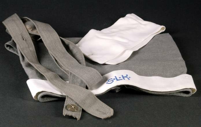 Klänning av grått ylletyg. Knälång klänning med isydd lång ärm. Kjolen har två nedsydda vecka fram och två fickor och ett veck bak. Knäppning från krage till midja med tio grå knappar.Knappar i handled.  Påsydda axelklaffar. Manchetter i vitt, löstagbara, påknäppt vit krage av konstsilke.Till klänningen hör skärp med knappar och knapphål och huvudbonad av samma tyg, snibb med knappar bak, påknäppt itt band fram med blått broderi: S.L.K (Sveriges lottakår). Mössan är märkt med samma UM nummer som klänningen.  Klänningen har löstagbara märken på axelklaffarna : 2. På bröstet  flera märken se UM 25.916. Ärmlängd 48 cm, axelbredd 48 cm, vidd 200 cm.