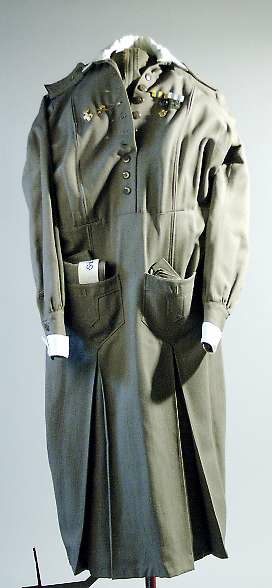 Klänning av grått ylletyg. Knälång klänning med isydd lång ärm. Kjolen har två nedsydda vecka fram och två fickor och ett veck bak. Knäppning från krage till midja med tio grå knappar.Knappar i handled.  Påsydda axelklaffar. Manchetter i vitt, löstagbara, påknäppt vit krage av konstsilke.Till klänningen hör skärp med knappar och knapphål och huvudbonad av samma tyg, snibb med knappar bak, påknäppt itt band fram med blått broderi: S.L.K (Sveriges lottakår). Mössan är märkt med samma UM nummer som klänningen.  Klänningen har löstagbara märken på axelklaffarna : 2. På bröstet  flera märken se UM 25.916. Ärmlängd 48 cm, axelbredd 48 cm, vidd 200 cm.