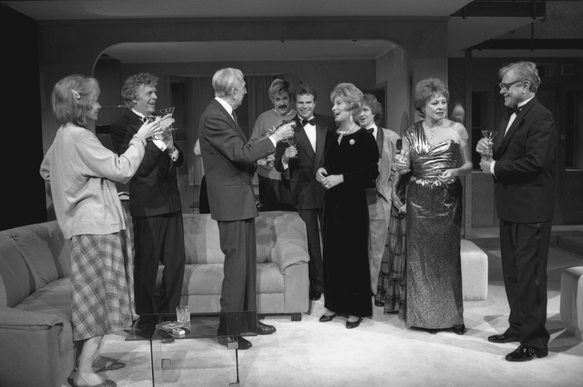 Scene fra Nationaltheaterets oppsetning av Alan Ayckbourns "Monki Bisniss". Elleve kvinner og menn kledd for fest står i en gruppe, der de prater og drikker. I forgrunnen sitter en ung mann og leser en bok.  Forestillingen hadde premiere 8. september 1989. Toralv Maurstad hadde regi og medvirkende var blant annet Tor Stokke som Jack, Kari Simonsen som Poppy og Leif Juster som Ken Ayres. 