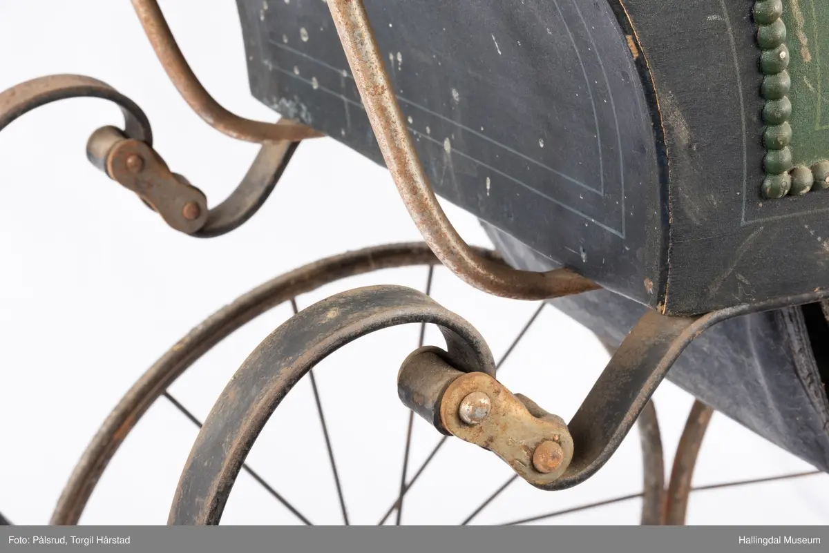 En barnevogn med jernunderstell/ramme, finérkasse trekt innvendig med svart lær (lakkert) og håndtak i messing og porselen. To sider har grønn, pålimt pyntelist rundt grønt felt omkranset av en svart kant. Påmalt, lys linjebord rundt grønt og svart felt på alle fire sidene. Setet kan justeres i sittende posisjon. Nedfellbar bunn. Fire hjul. Kalesje mangler.
