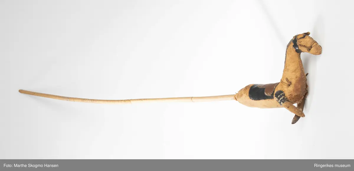 En kjepphest med lang, tynn pinne. Foran er det ikke et hode, men en hel forkropp av en hest. Hesten er laget av lær, som er godt stoppet. Hver side av kropp, hals og hode er laget av ett stykke lær. De to lærstykkene er sydd sammen langs rygg og buk. Salen er laget av to stykker lær som er sydd fast oppå dette igjen. Manen er laget ved at ull er sydd på tvers av nakken. Forbeina er proporsjonalt veldig små, og er sydd fast i kroppen med et stykke svart lær på toppen av beina. Rundt hodet er det et bånd av svart lær, ørene stikker fram fra under dette. Det er lærsnorer å tvers av mulen og sydd gjennom mulen der munnen skulle ha vært. Tilsvarende lærsnorer er sydd gjennom brystet og ligger over nedre del av nakke. Skal muligens illudere tøyler, men de er ikke komplette.
