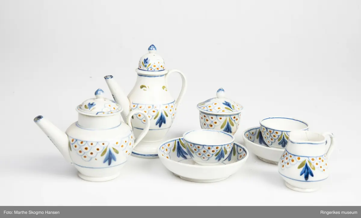 Dokkeserviset består av to skåler (G&H), to boller eller kopper (uten hank) (E&F), en fløtemugge (C), en sukkerkopp (D), en tekanne (B) og en kaffekanne (A). Kannene har tut og hanker, og lokk. Tuten på kaffekanna har vært brukket, og er limt på igjen. Sukkerkoppen har også lokk. Lokkene har små knopper som håndtak, disse er blå på toppen. Sukkerkoppen har ikke håndtak, men har lokk. Serviset er dekorert med blått, oransje, og grønt. Noen av dekorasjonene er blomsteraktige, mens andre er buer med prikker inni.