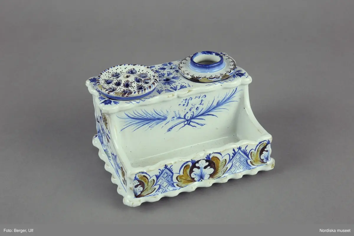 Katalogkort:
"Skrivtyg, fajans. Rektangulärt, framtill rum för pennor, upptill bläckhorn och sanddosa; dekor. med blommor och rutmönster i blått, brunt och gult, på framsidan två palmkvistar i blått, varöver H.H. 
Sign. i blått: Stockholm 2/8 1749 Hindrich Höök.
/Upmark 1904"

Skrivställ av fajans med dekor i blått och manganviolett, Rörstrand 1749.
Skrivstället med runda dosor för bläck och strösand är målat av Henrik Höök som signerat det med hela sitt namn under botten, oftast står bara målarens initialer utsatta. Han var då lärgosse vid fabriken och blev målargesäll 1754. 
Märkning: "Stockholm 2/8 1749" samt "Hindrich Höök".
Byggmäst E A Bomans samling nr  75.
/Annika Tegnér 2006