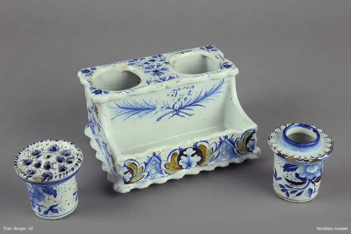 Katalogkort:
"Skrivtyg, fajans. Rektangulärt, framtill rum för pennor, upptill bläckhorn och sanddosa; dekor. med blommor och rutmönster i blått, brunt och gult, på framsidan två palmkvistar i blått, varöver H.H. 
Sign. i blått: Stockholm 2/8 1749 Hindrich Höök.
/Upmark 1904"

Skrivställ av fajans med dekor i blått och manganviolett, Rörstrand 1749.
Skrivstället med runda dosor för bläck och strösand är målat av Henrik Höök som signerat det med hela sitt namn under botten, oftast står bara målarens initialer utsatta. Han var då lärgosse vid fabriken och blev målargesäll 1754. 
Märkning: "Stockholm 2/8 1749" samt "Hindrich Höök".
Byggmäst E A Bomans samling nr  75.
/Annika Tegnér 2006