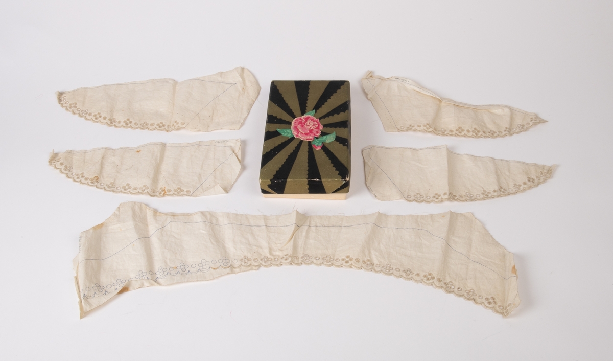 Krave og mansjetter, uferdige, engelsk broderi.  
a) Krave. Et stykke stoff med inntegnet  krave med blåblyant, vel 2/3 av kanten er ferdig  brodert.  
b-e) 4 stk. trekantede mansjetter, ferdig broderte,  men ikke klippet etter mønsterstrekene.