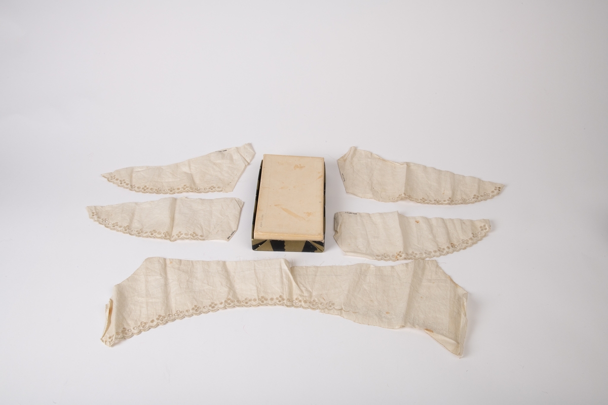 Krave og mansjetter, uferdige, engelsk broderi.  
a) Krave. Et stykke stoff med inntegnet  krave med blåblyant, vel 2/3 av kanten er ferdig  brodert.  
b-e) 4 stk. trekantede mansjetter, ferdig broderte,  men ikke klippet etter mønsterstrekene.