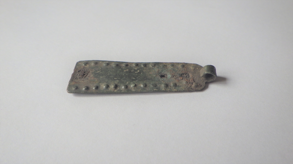 Rektangulært og trapesformet beslag som ender i et smalt bånd i den smaleste enden. Båndet er bøyd rundt slik at det utgjør en ring i enden av beslaget. Gjenstanden er dekorert med innpunslede perler i rad langs alle kanter. Beslaget har tegn til tre naglehull, hvorav to fortsatt har rester av nagler sittende i.