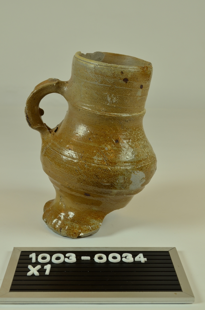 Siegburgkeramikk. Steingods. Grålig gods. Lysebrun glasur. Med hank. Del av stett og bunn avslått. Ytre diameter munning 7. Stett med buklete kant. Diam stett 8,9. :: Dateringsreferanse katalognummer 503 i Keramikk 1000-1600 tyder på 1400-tallet.