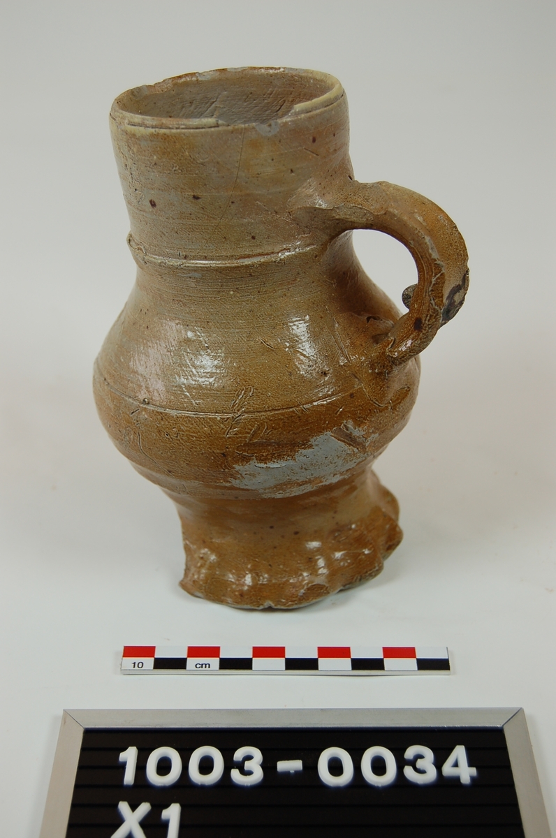 Siegburgkeramikk. Steingods. Grålig gods. Lysebrun glasur. Med hank. Del av stett og bunn avslått. Ytre diameter munning 7. Stett med buklete kant. Diam stett 8,9. :: Dateringsreferanse katalognummer 503 i Keramikk 1000-1600 tyder på 1400-tallet.