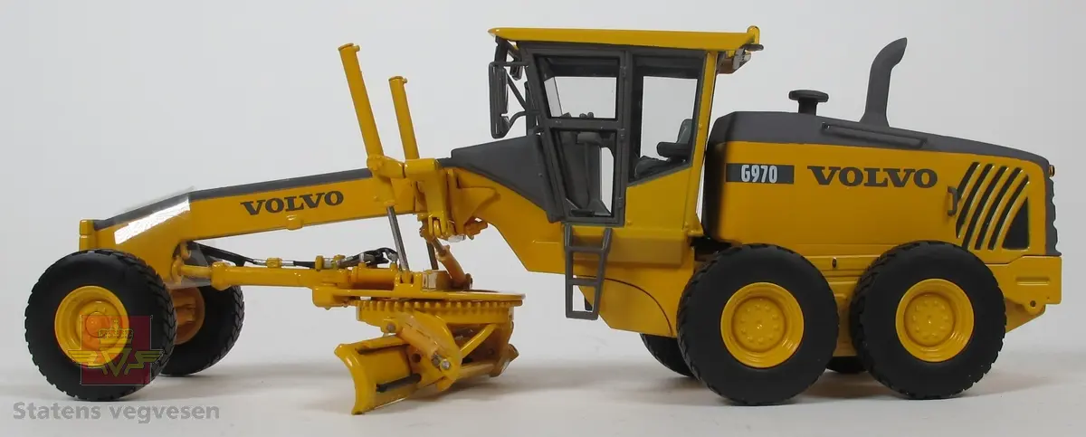 Volvo G970. Miniatyrmodell av veghøvel i eske. Esken har fargene svart, grå og burgunder samt påskriftene MOTORART collectible models og VOLVO. Den har også en skade i et av hjørnene der pappen har revnet. Modellen er laget hovedsakelig i metall med deler i plast og gummi. Fargen er gul med detaljer i svart og grått. Veghøvelen har tre akslinger, (seks hjul), og skjær. Merking fra produsenten under veghøvelen samt Volvo G970 merking utenpå. 
Skala 1:50.