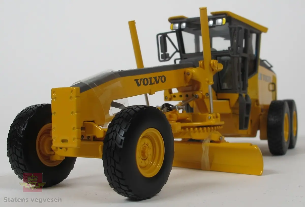 Volvo G970. Miniatyrmodell av veghøvel i eske. Esken har fargene svart, grå og burgunder samt påskriftene MOTORART collectible models og VOLVO. Den har også en skade i et av hjørnene der pappen har revnet. Modellen er laget hovedsakelig i metall med deler i plast og gummi. Fargen er gul med detaljer i svart og grått. Veghøvelen har tre akslinger, (seks hjul), og skjær. Merking fra produsenten under veghøvelen samt Volvo G970 merking utenpå. 
Skala 1:50.