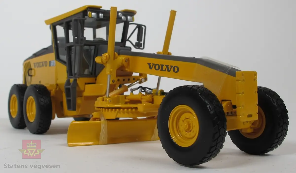 Volvo G970. Miniatyrmodell av veghøvel i eske. Esken har fargene svart, grå og burgunder samt påskriftene MOTORART collectible models og VOLVO. Den har også en skade i et av hjørnene der pappen har revnet. Modellen er laget hovedsakelig i metall med deler i plast og gummi. Fargen er gul med detaljer i svart og grått. Veghøvelen har tre akslinger, (seks hjul), og skjær. Merking fra produsenten under veghøvelen samt Volvo G970 merking utenpå. 
Skala 1:50.